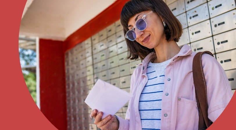 A woman in sunglasses standing at a row of community mailboxes, reading an envelope she has just collected.