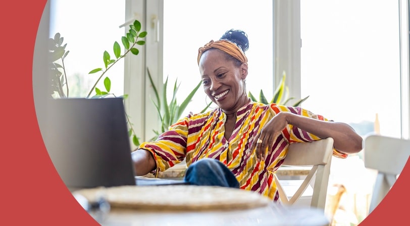 A smiling woman relaxing at a sunlit table at home while browsing on her laptop.