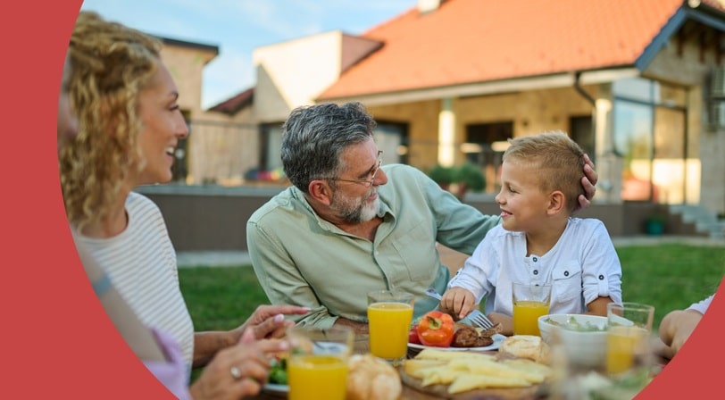 A multigenerational family shares an outdoor lunch in the backyard as a grandfather smiles at his grandson.
