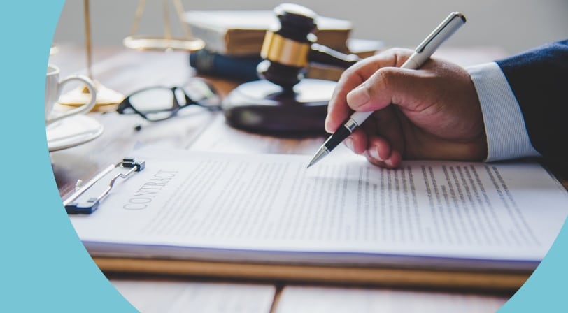 A hand holding a pen over a printed contract on a wooden desk, with a gavel, scales, and reading glasses nearby.