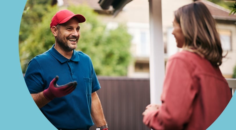 A maintenance worker in a red cap and tool belt smiling as he speaks with a resident on her front porch.