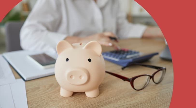 A piggy bank on a desk in the foreground while a person works through paperwork with a calculator behind it.