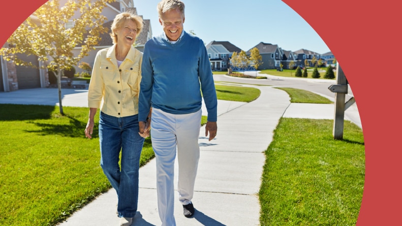 A smiling older couple holding hands on a sidewalk stroll through their 55+ community neighborhood.