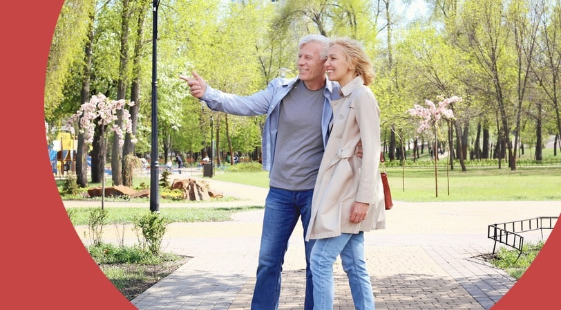 A 55+ couple walking in a park, one pointing at something in the distance, with spring trees in bloom.