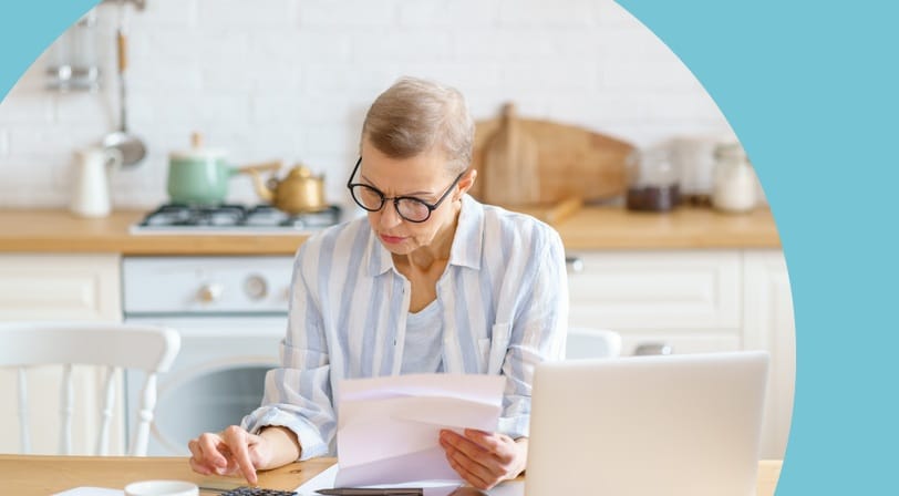 An older woman sitting at her kitchen table with a laptop and calculator, reviewing paperwork.
