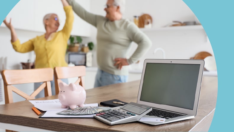 A laptop, calculator, cash, and refund form on a table while a happy 55+ couple dance in the kitchen.