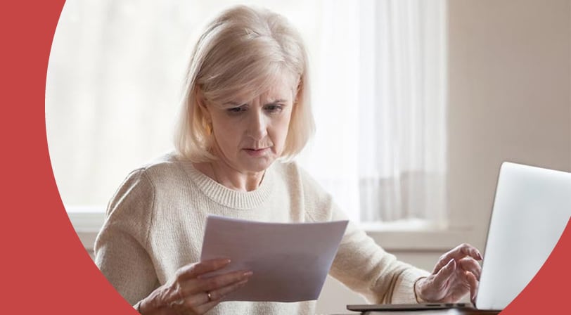 A woman with light blonde hair sits at a wooden desk reading a printed document with a concerned expression, one hand resting on an open laptop.