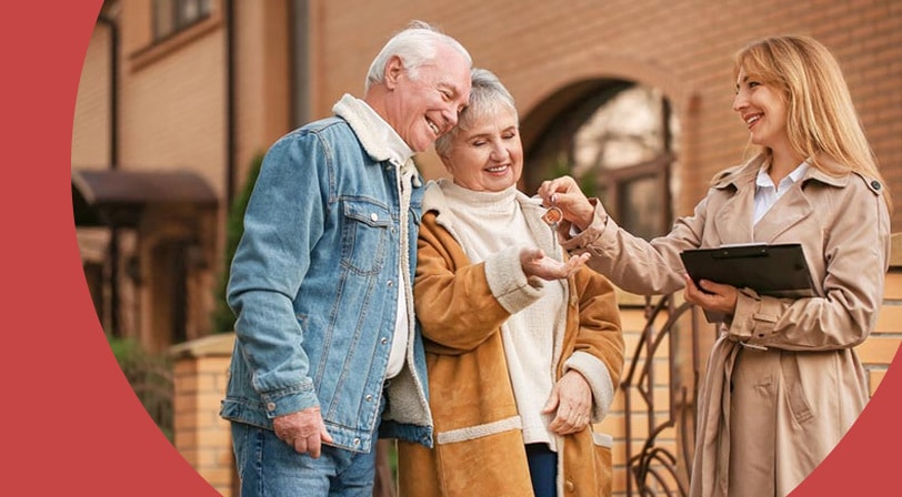 A smiling couple receives a set of house keys from a real estate agent holding a clipboard outside a brick home.