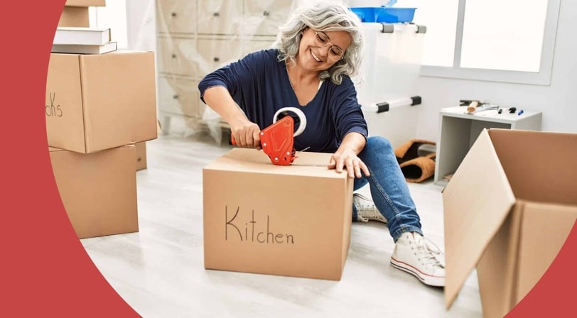 Woman taping a cardboard box labeled "Kitchen" while sitting on the floor of a nearly empty room surrounded by moving boxes.