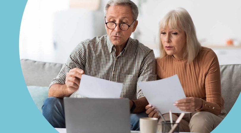 Senior couple reviewing printed documents together on a couch with a laptop and coffee mugs on the table in front of them.