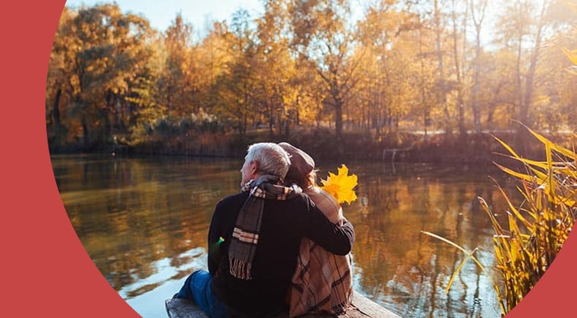 A 55+ couple sitting on a dock overlooking a lake surrounded by autumn foliage.