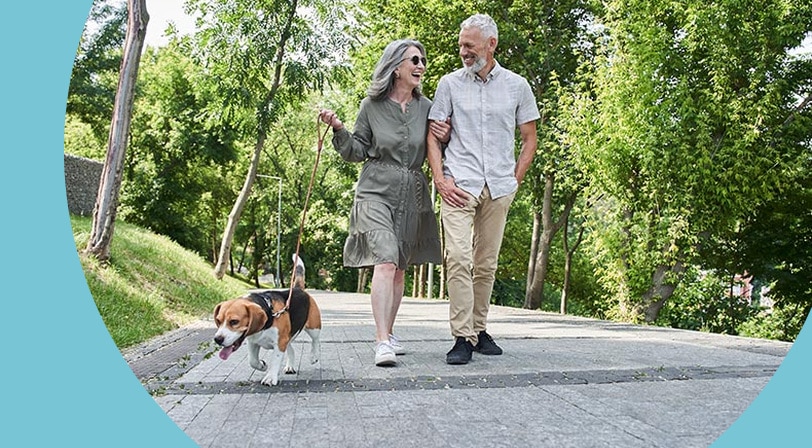 A 55+ couple walking their beagle along a paved park path on a summer day.