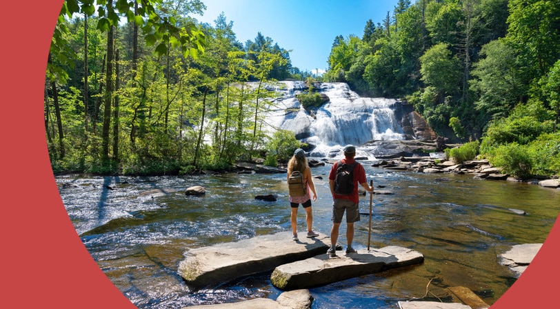 Two hikers standing on flat rocks at the edge of a river, looking up at a wide cascading waterfall surrounded by forest.