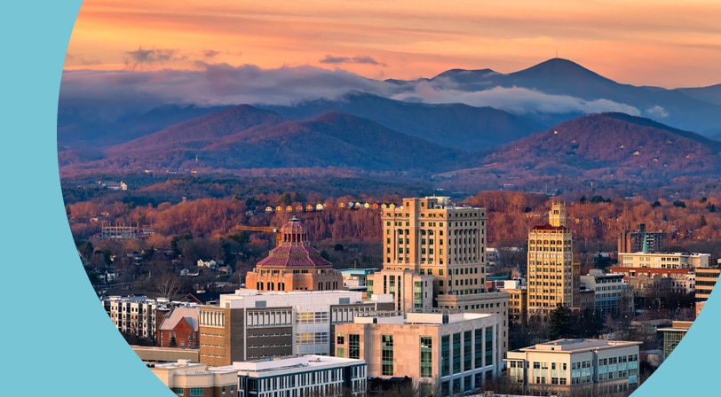 Aerial view of downtown Asheville, North Carolina, with historic high-rise buildings in the foreground and the Blue Ridge Mountains under an orange sunset sky.