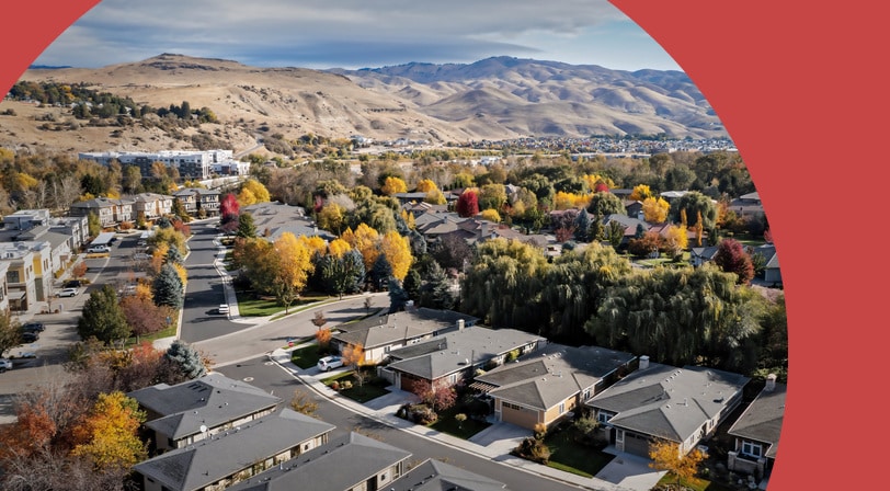 An aerial view of a Boise neighborhood of single-story homes and tree-lined streets in fall color, with the foothills rising behind it.