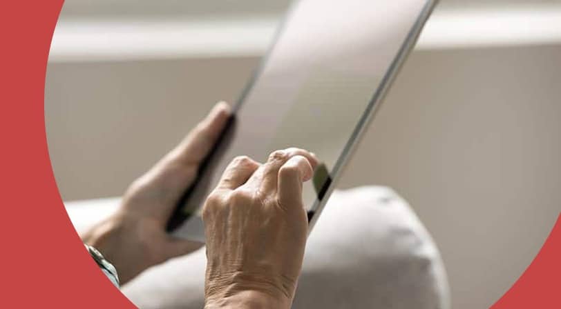 An older woman’s hands holding a tablet on a light gray sofa, with one finger touching the screen.
