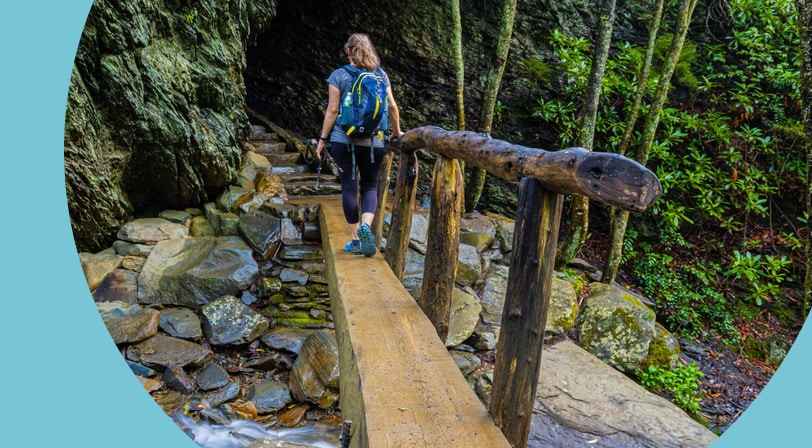 A hiker with a backpack crossing a narrow wooden footbridge toward a mossy rock overhang on a forest trail.