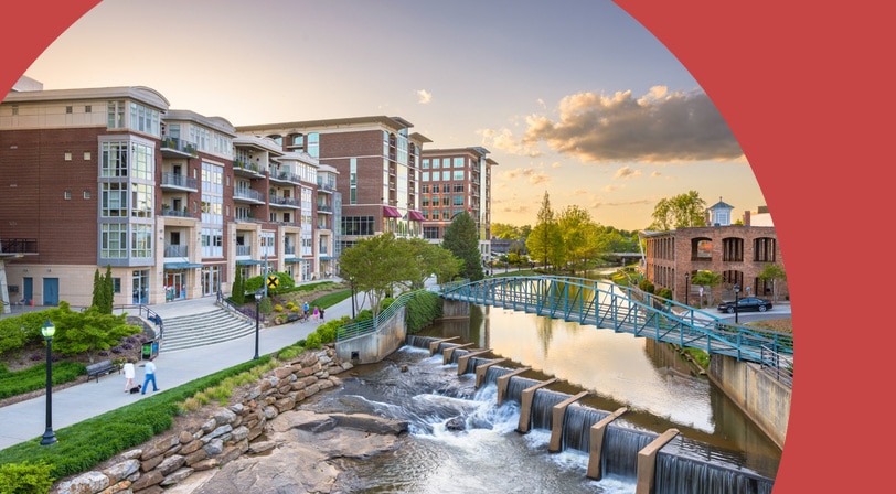 The Reedy River falls and a blue pedestrian bridge in downtown Greenville, South Carolina, lined with brick apartment buildings and riverside walking paths at sunset.