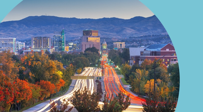 The downtown Boise skyline at dusk, with the Idaho State Capitol dome lit at the end of a tree-lined boulevard and car light trails streaking along the road.