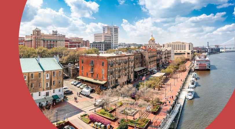 Aerial view of Savannah, Georgia’s River Street waterfront, with historic brick warehouses, a landscaped riverside plaza, and a paddlewheel riverboat docked on the Savannah River.