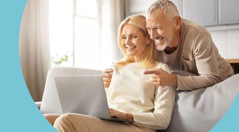 A smiling couple looking at a laptop together, the woman seated on a sofa and the man leaning over the back beside her, in a bright living room.