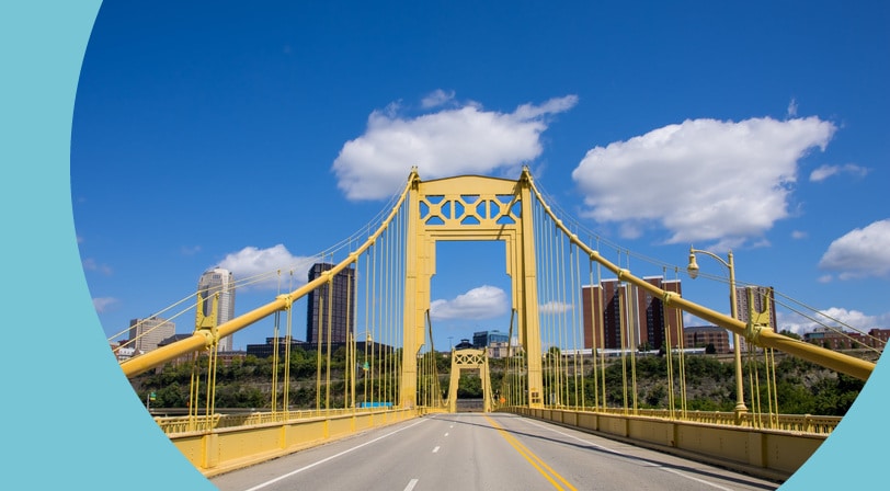 The yellow suspension towers of a Pittsburgh bridge over the Monongahela River, with the city skyline and a bright blue sky in the background.