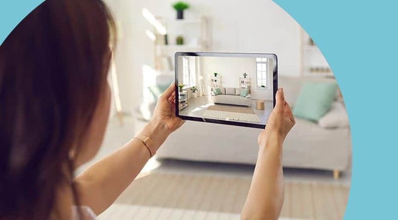A woman holding up a tablet that displays a bright, modern living room with a sofa, shelves, and houseplants.