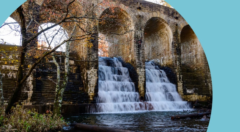 Water cascading over a stone arch dam and bridge at Cumberland Mountain State Park near Crossville, Tennessee, surrounded by autumn foliage.