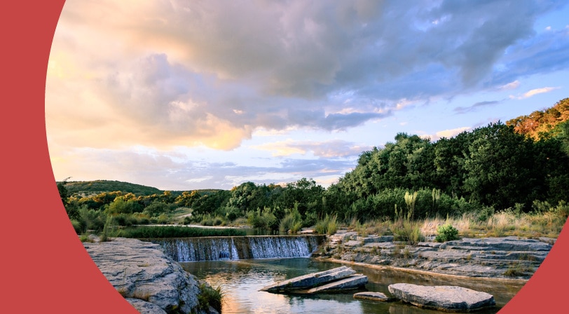 A low waterfall spilling over a limestone ledge into a calm pool, with wooded hills and a pastel sunset sky behind it.