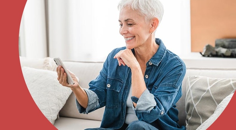 A smiling woman with short silver hair sitting on a cream sofa and looking at her smartphone.