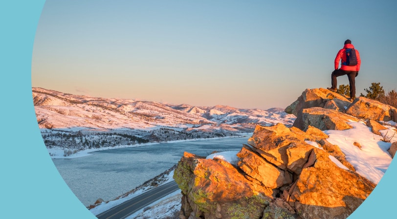 A hiker in a red jacket standing on a rocky outcrop above a reservoir and snow-covered foothills at sunrise.