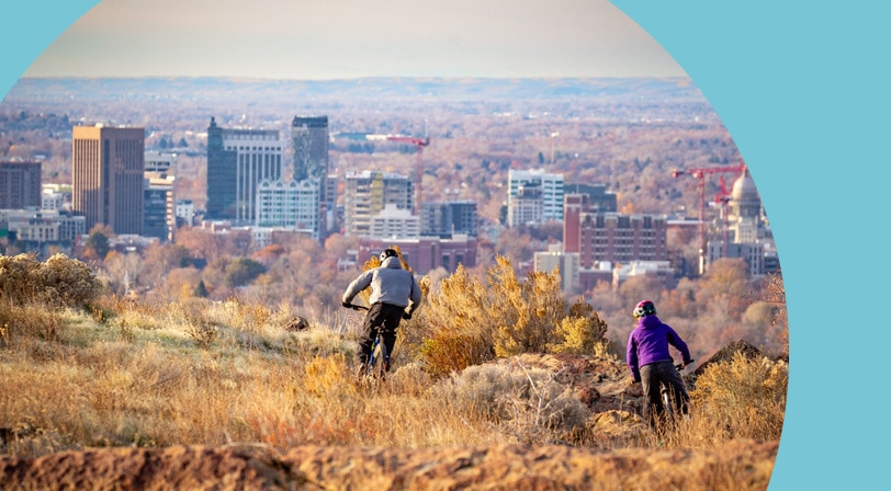 Two mountain bikers riding a foothills trail through dry golden grass with the Boise skyline spread out below them.