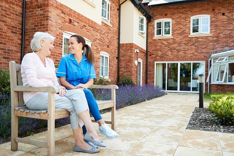 A senior woman sitting on a bench and talking with a nurse in retirement home.