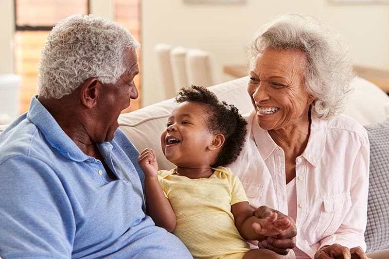 Grandparents sitting on sofa at home playing with baby granddaughter.