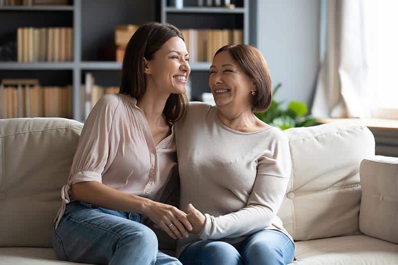 A 55+ mother hugging her grown daughter and sitting on a cozy couch at home.
